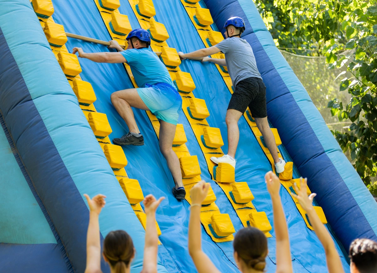 Two competitors race to the top of an inflatable climbing wall, surrounded by an enthusiastic crowd cheering them on as they tackle the colorful challenge.